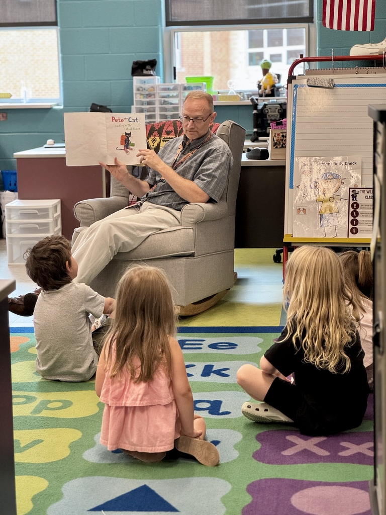 Students sit on carpet in front of a teacher who is reading a book