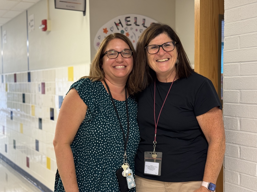 Two teachers smiling, standing outside of a classroom.