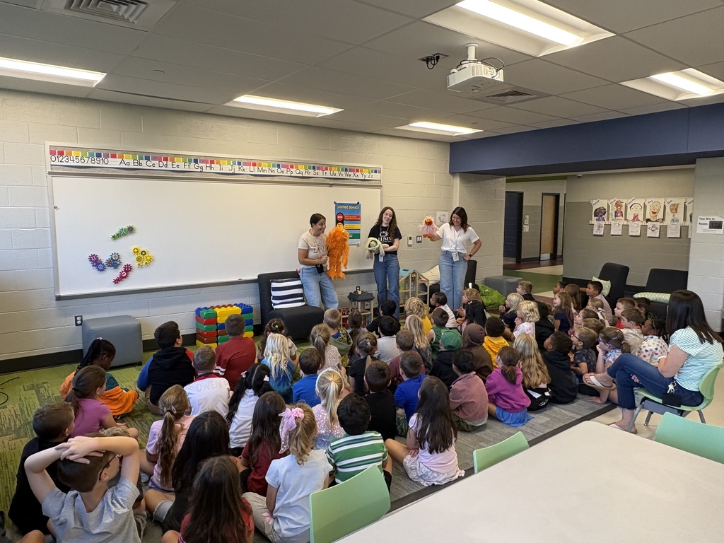A group of students sitting on a floor while teachers lead a lesson at a white board.