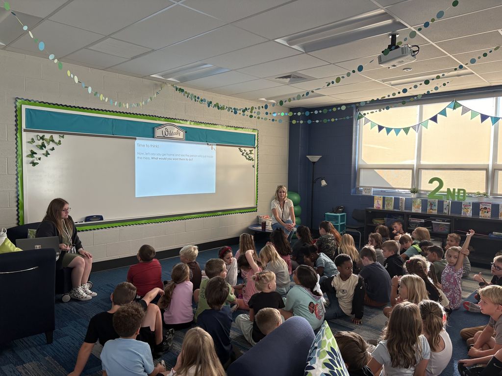 A group of students sitting on a floor while teachers lead a lesson at a white board.