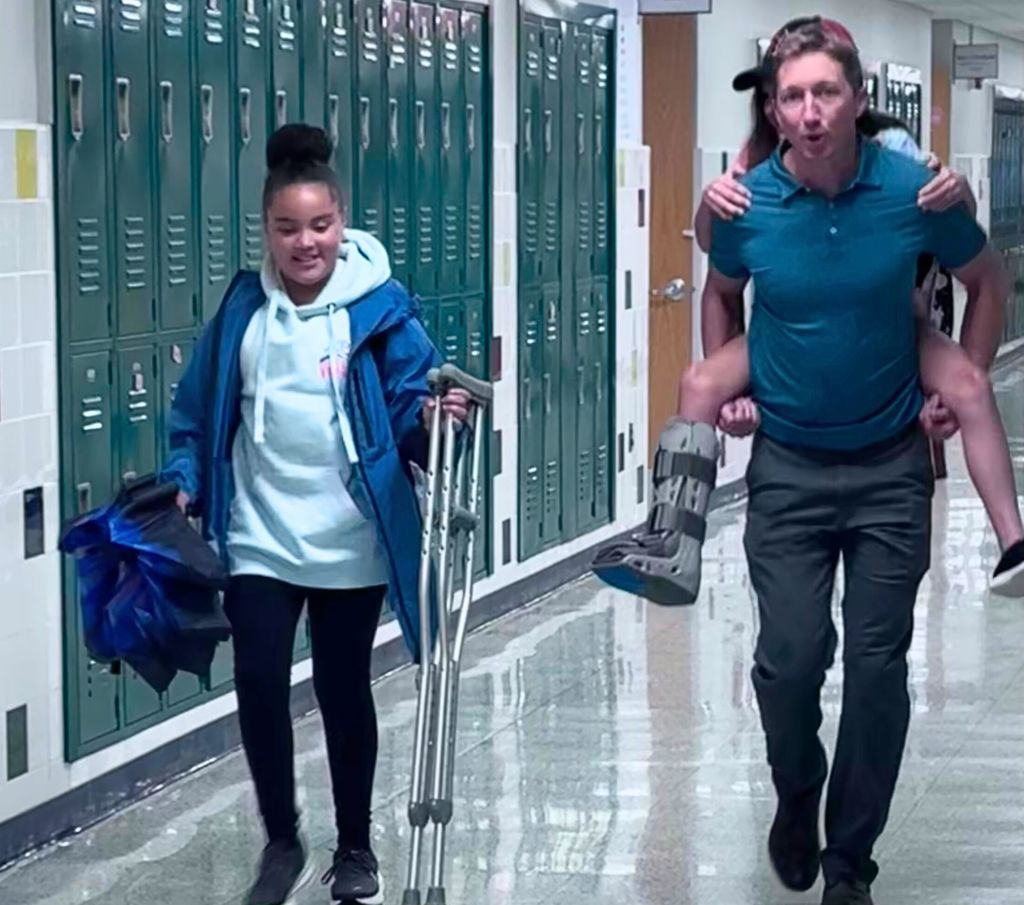 A teacher carrying a student on his back while another carries crutches and an umbrella.