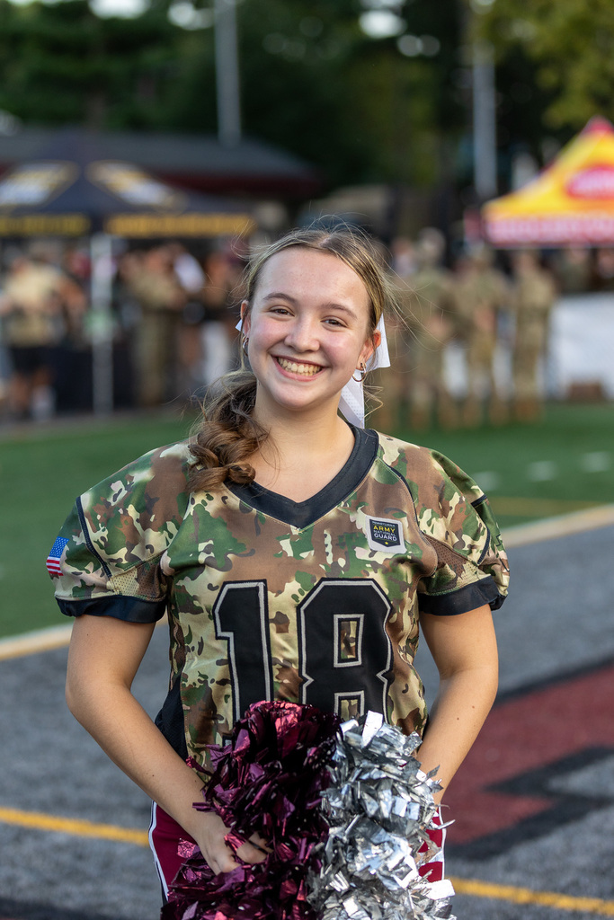 Cheerleader on sidelines of football game wearing a camo shirt and holding pom poms.