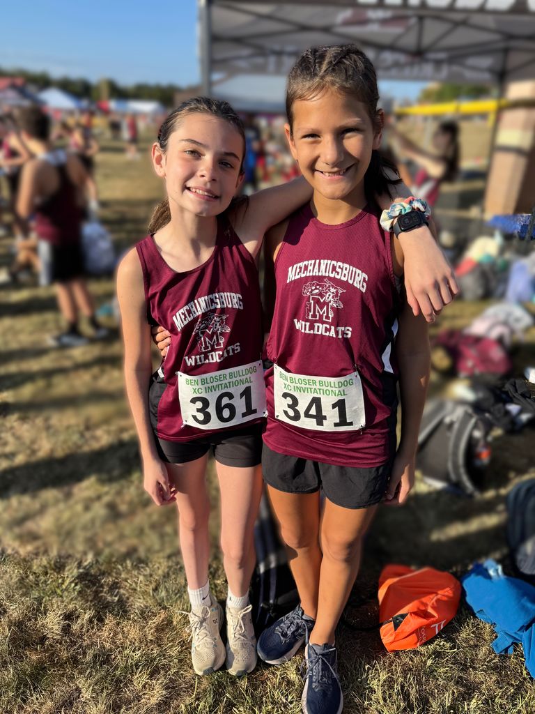 Two girls standing beside each other in maroon tank tops as they prep to run a XC meet
