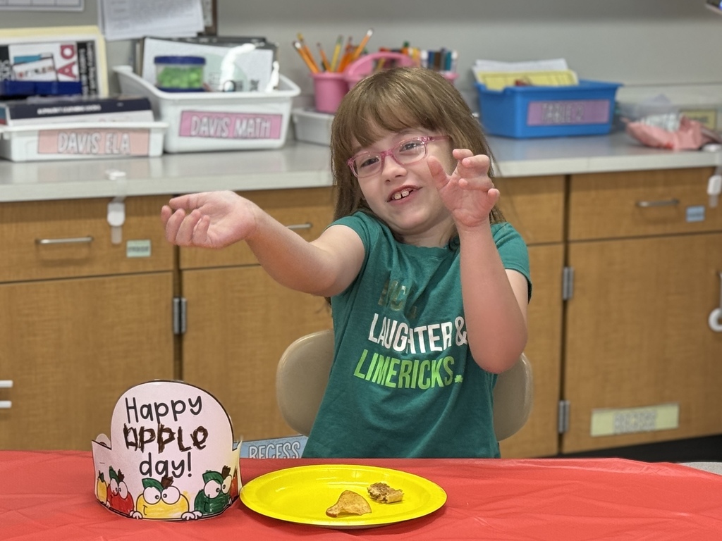 A student sits at a table wearing a Happy Apple Day hat and tastetesting apple foods.
