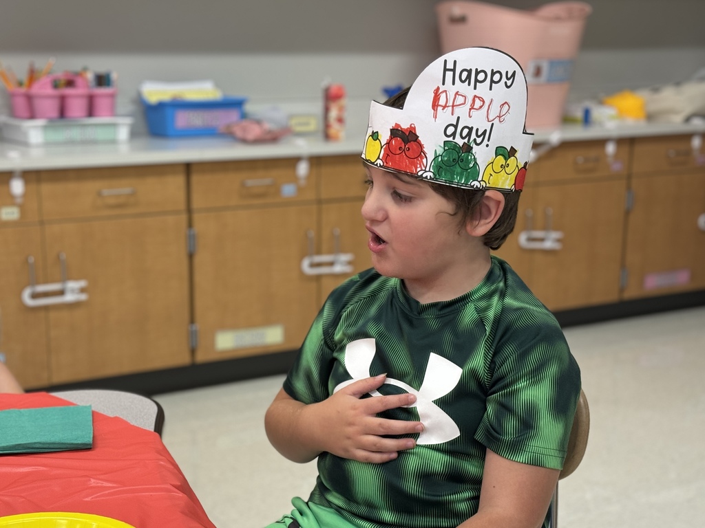 A student sits at a table wearing a Happy Apple Day hat and taste testing apple foods.