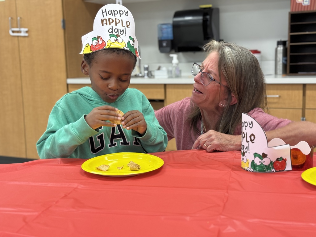 A student sits at a table wearing a Happy Apple Day hat and taste testing apple foods.