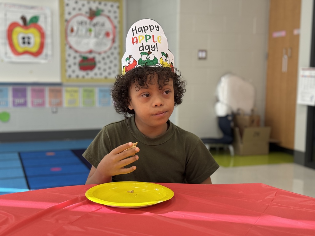 A student sits at a table wearing a Happy Apple Day hat and tastetesting apple foods.