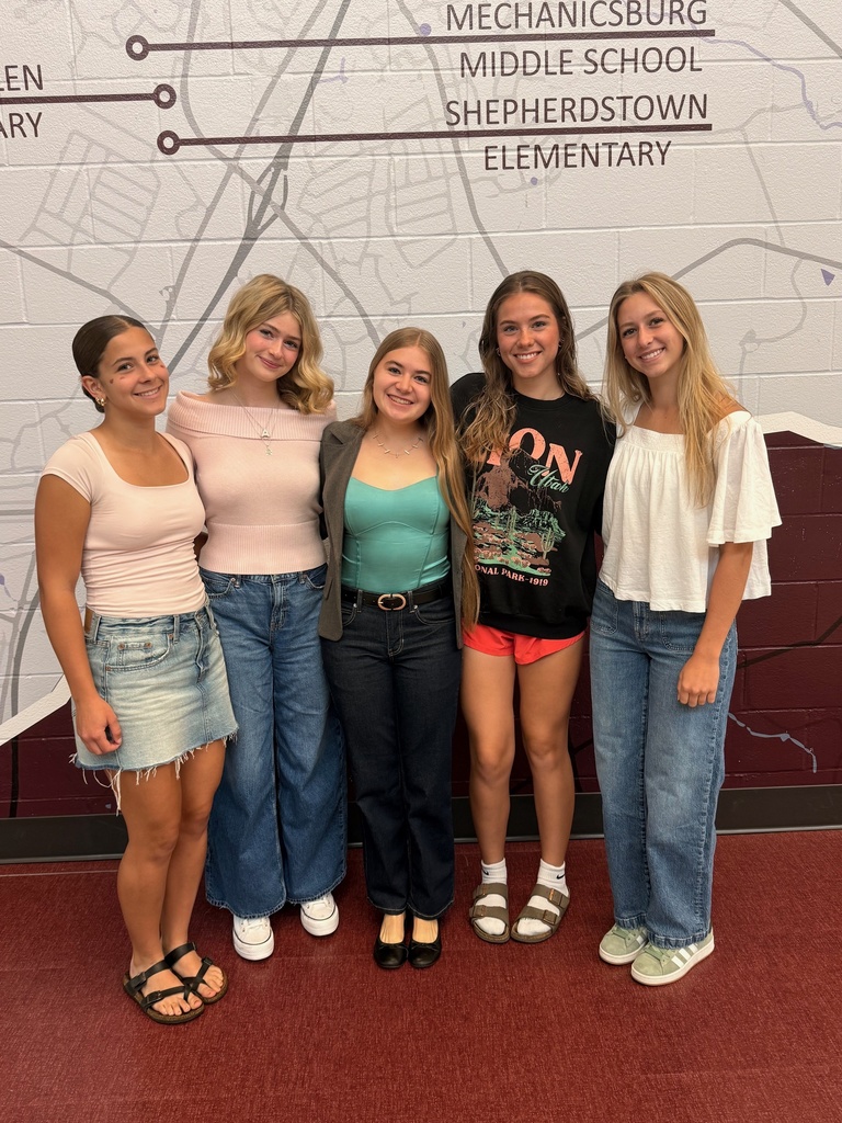 A group of five young ladies standing next to each other in front of a painted cement wall.
