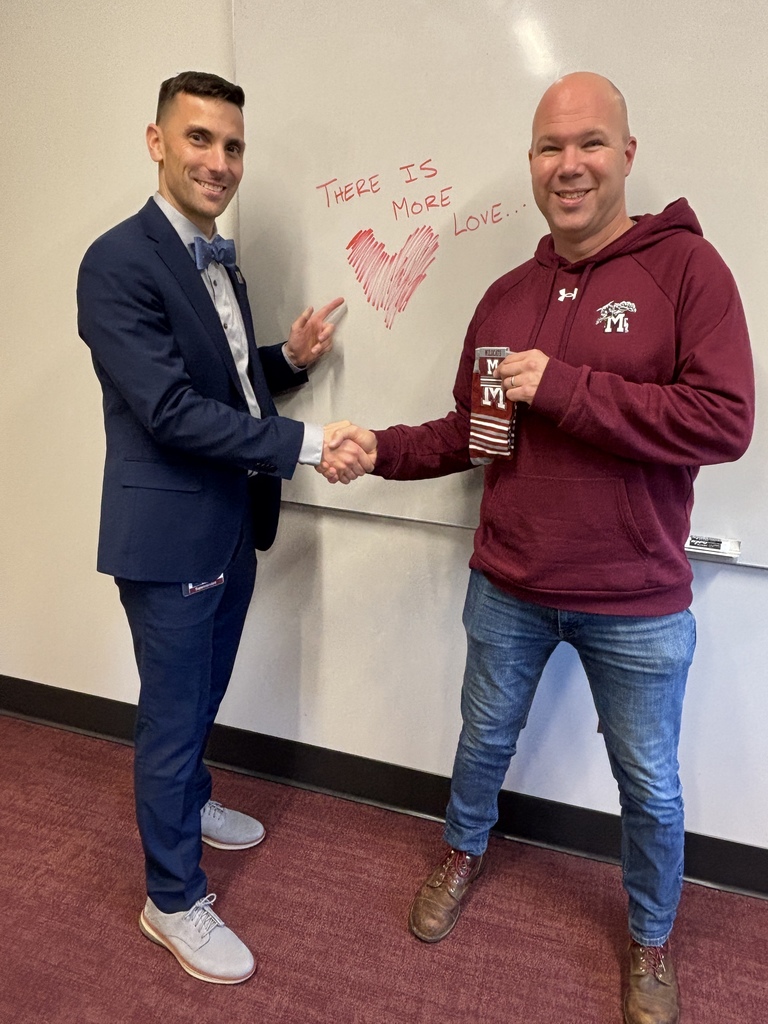 Two men shaking hands while holding a pair of branded socks and standing in front of a whiteboard that says  "There is More Love ..."
