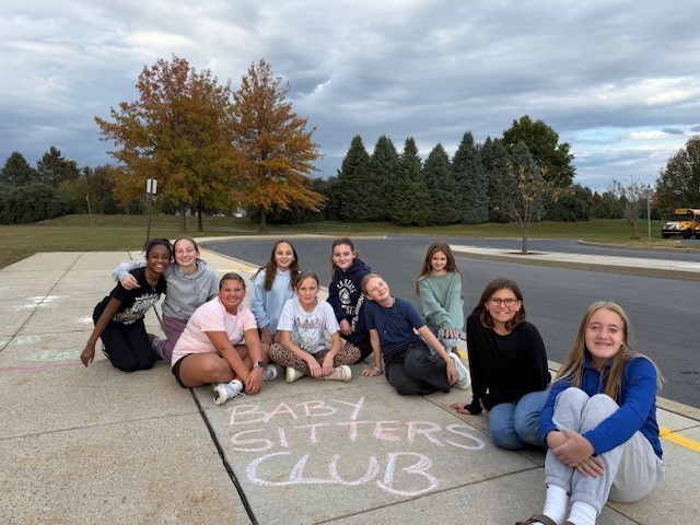 Students sitting outside on a sidewalk, where they wrote Babysitters Club in chalk.