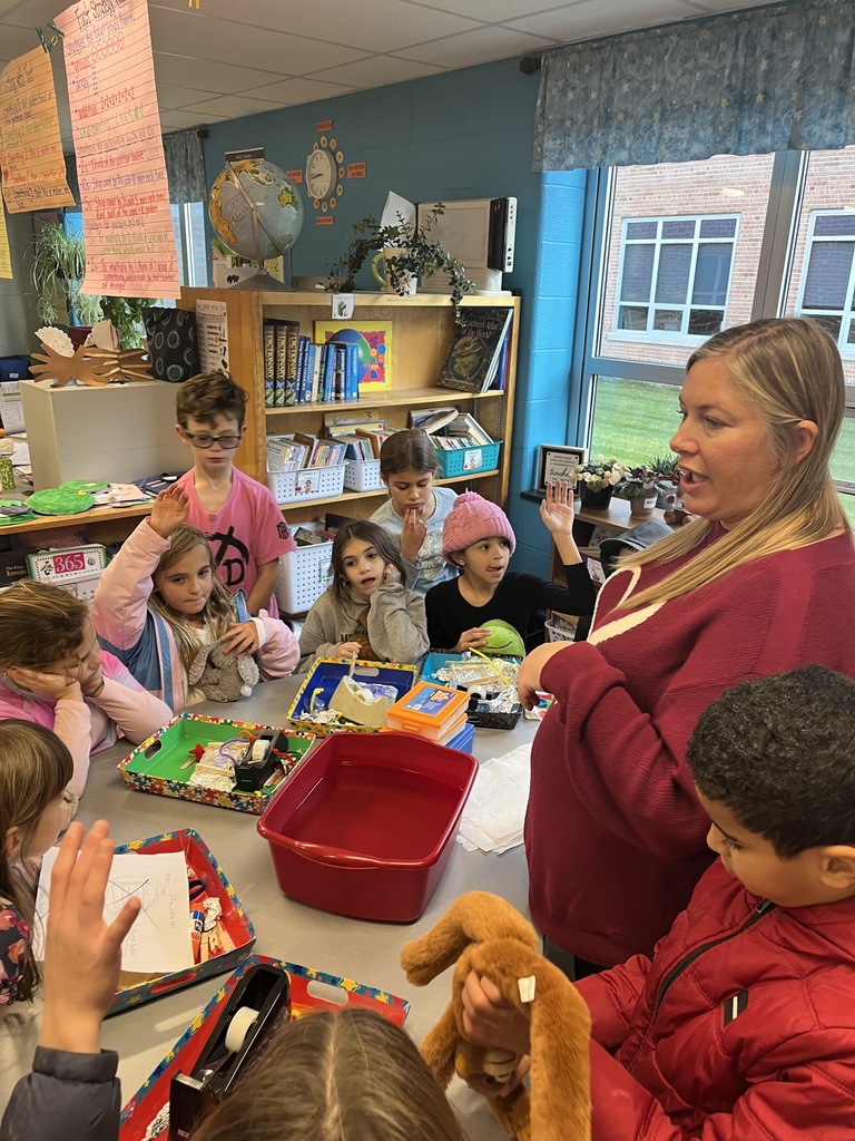 Students gathered around a table with items and a bucket of water, making predictions about what will sink and what will float.
