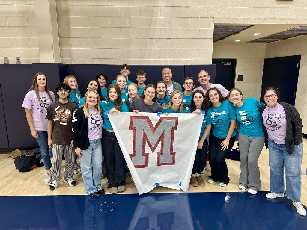 A group of students and staff gathered around a banner with a letter M.