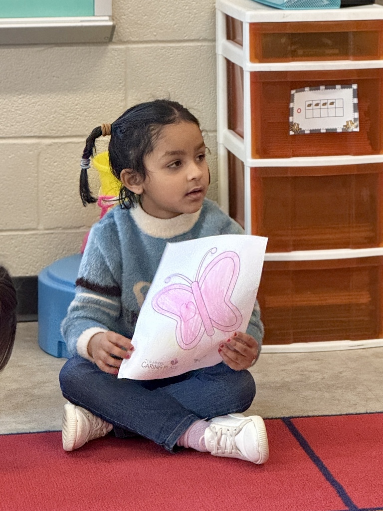 A young child sitting in circle time in school, holding a picture of a butterfly they colored.