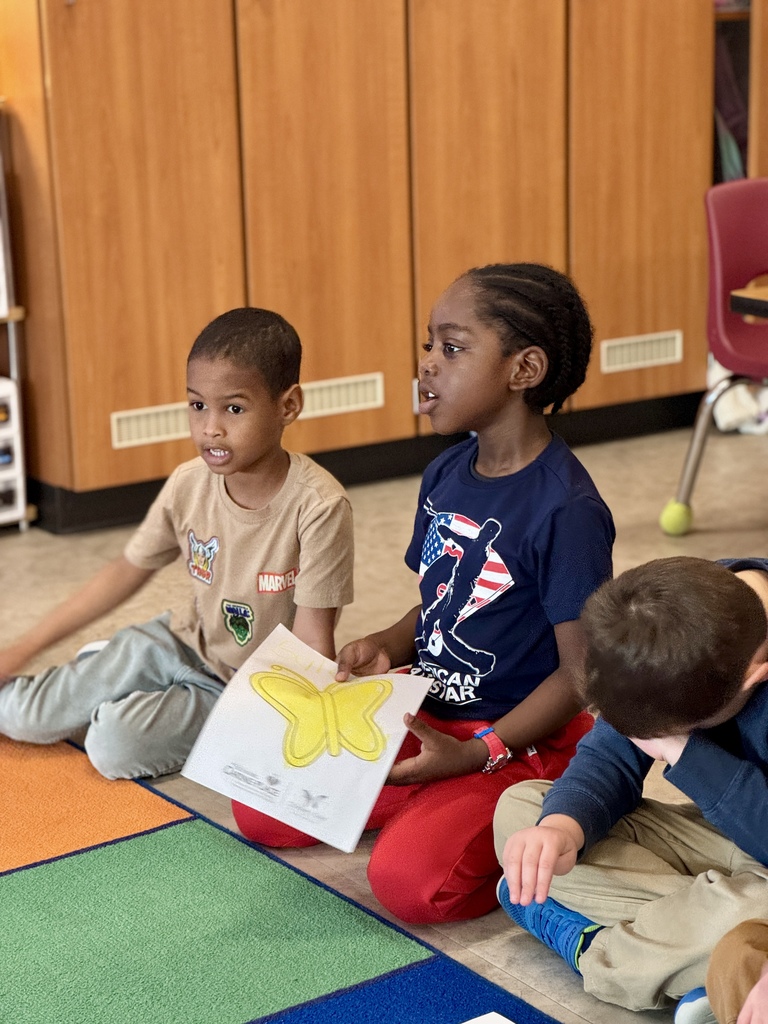 A young child sitting in circle time in school, holding a picture of a butterfly they colored.
