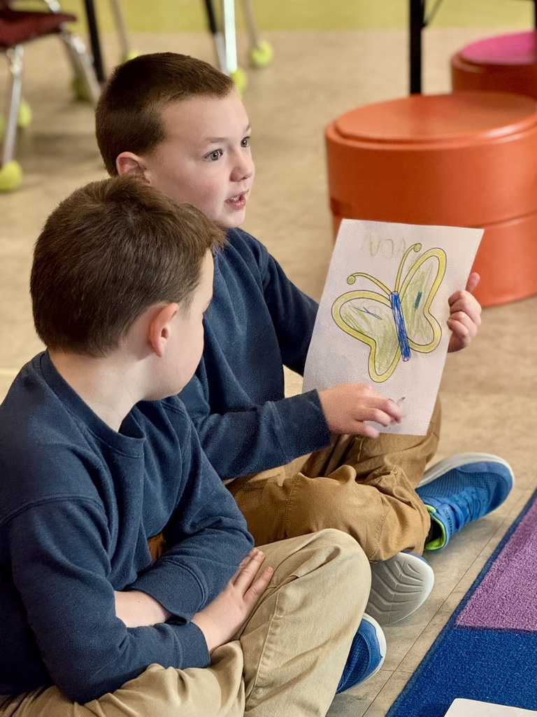 A young child sitting in circle time in school, holding a picture of a butterfly they colored.