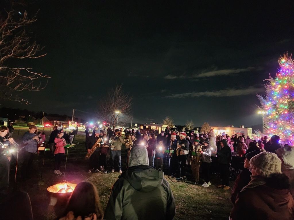 Marching band members performing around a campfire while guests look on.