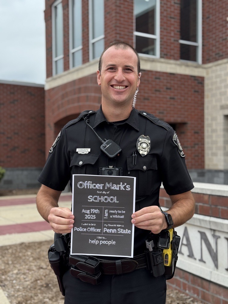 A police officer stands in front of a school and holds a first day of school sign.