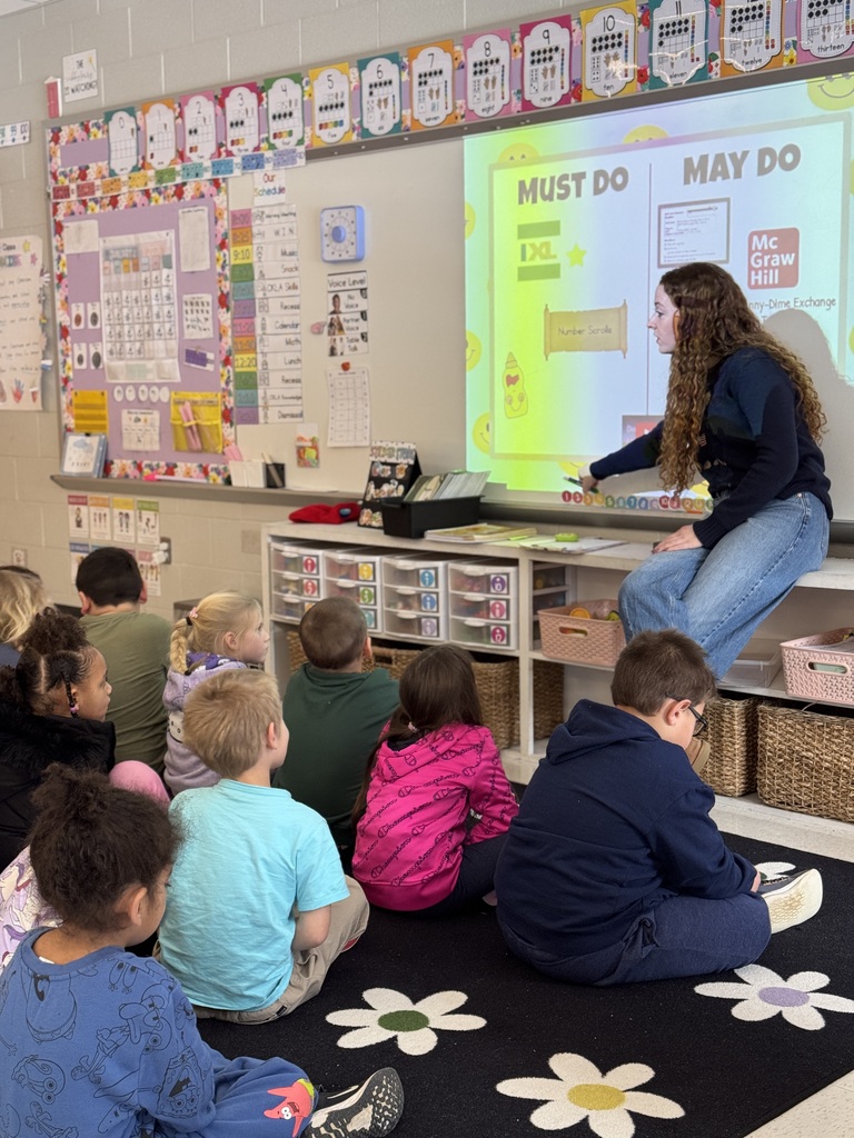 Students sit on the floor while their teacher displays instructions on a whiteboard.