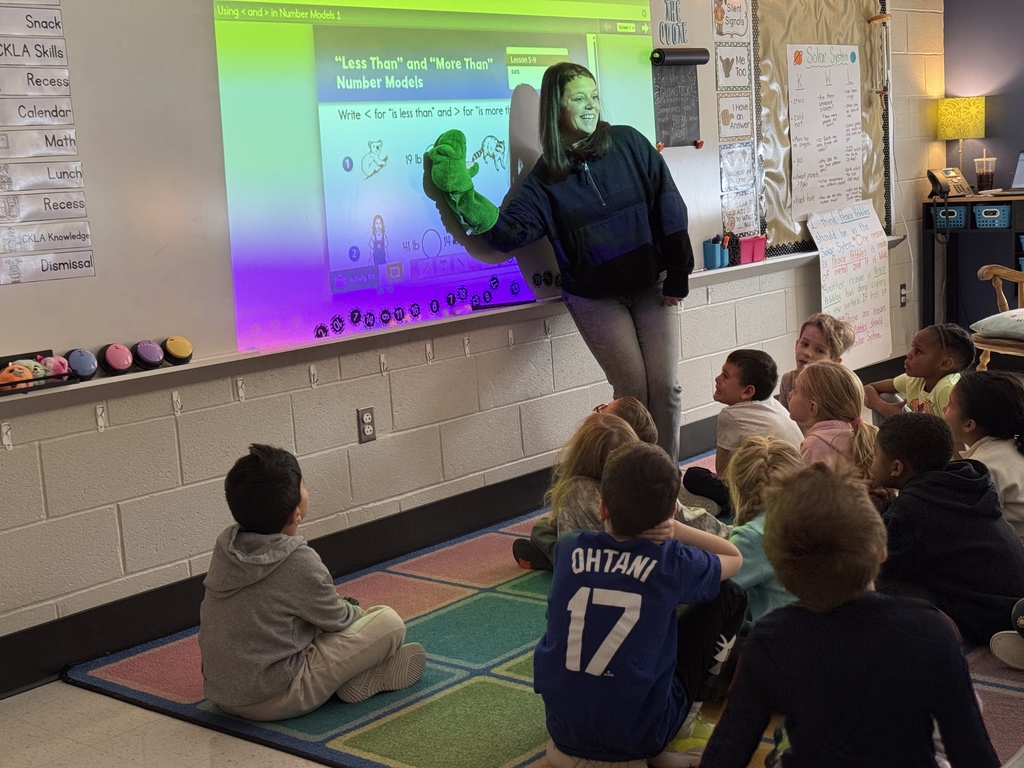 A teacher holding a puppet alligator as part of a lesson about greater and less than math.