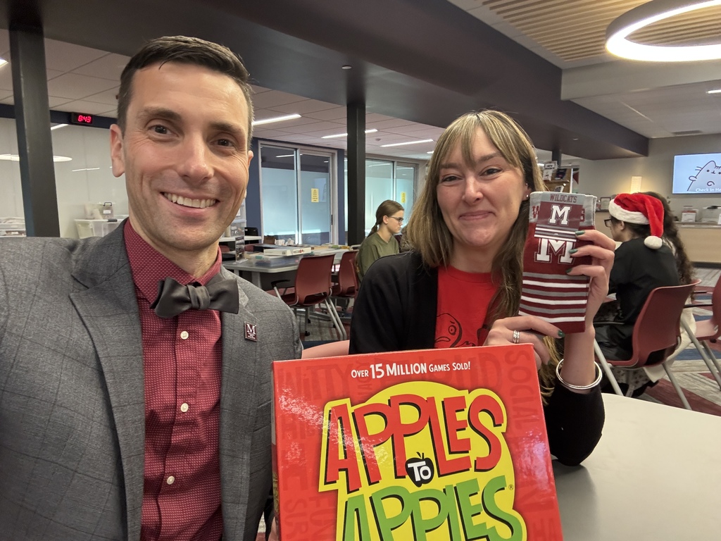 Two adult staff members holding up an Apples to Apples board game in a school library.