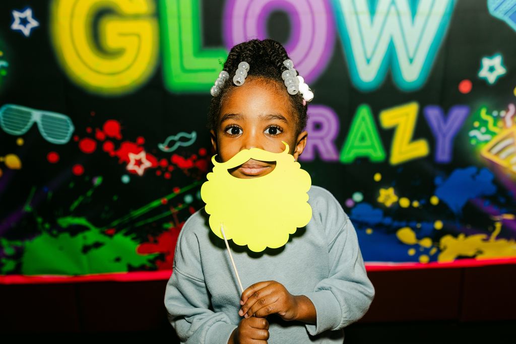 A young student at a glow party holding up a pretend beard.