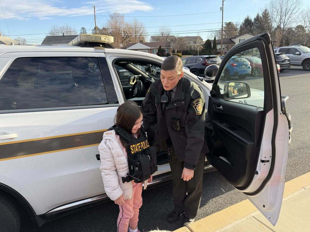A young students puts on a state police trooper's vest while the trooper looks on.
