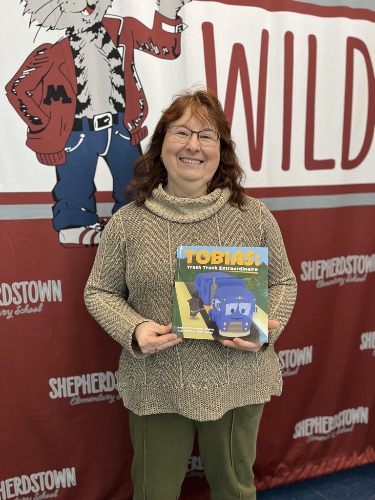 A teacher holds a book she wrote while standing in front of a maroon Wildcats banner.