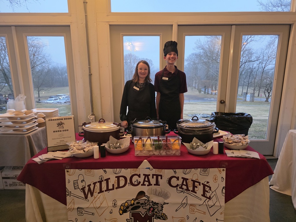 Two students at a community event, standing behind a table full of soups they made.