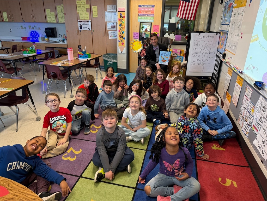 A group of young students sits on the floor and poses for a picture with a man in a suit who's holding a book.