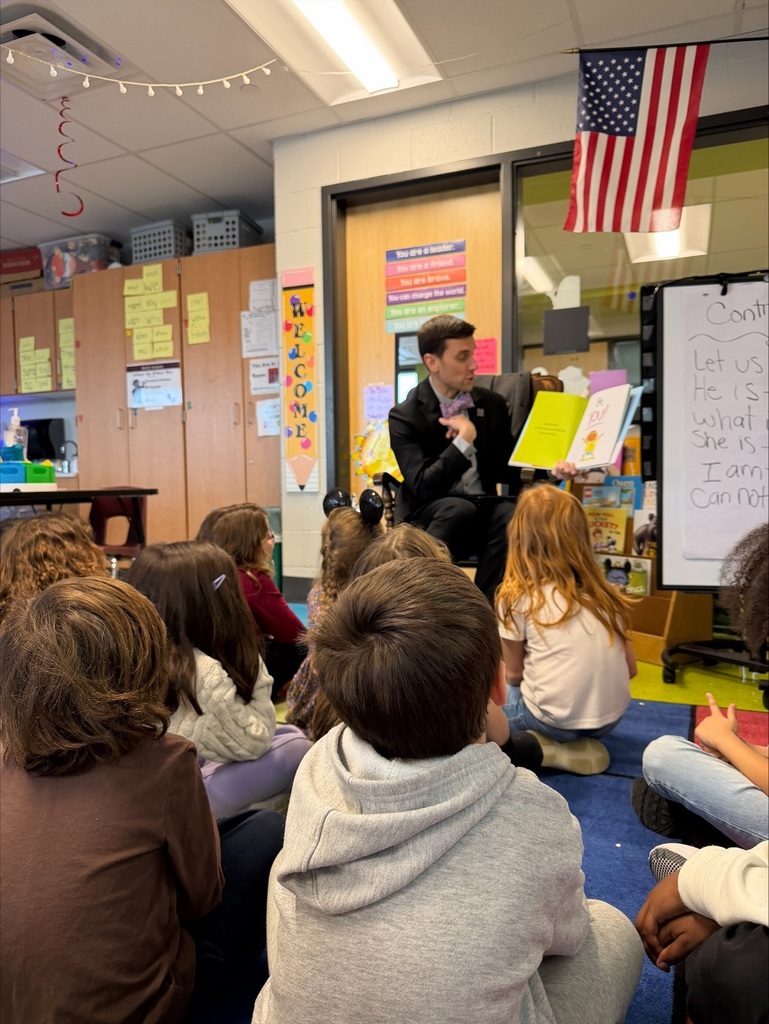 A man in a suit sits in a chair and reads a book to a group of elementary students.