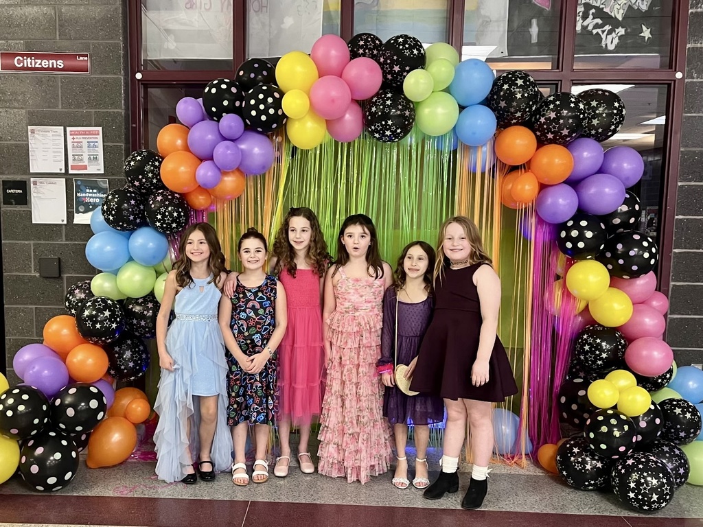 A group of young students dressed up and standing under a colorful balloon arch.