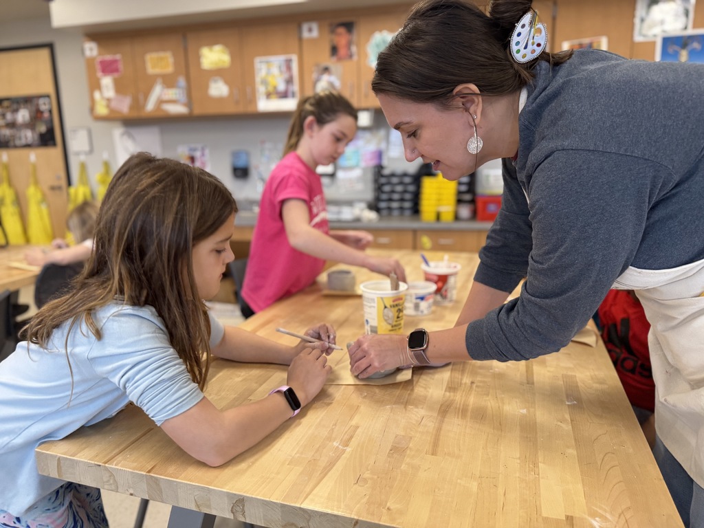 A teacher helps a young student shape a clay pot in art class.