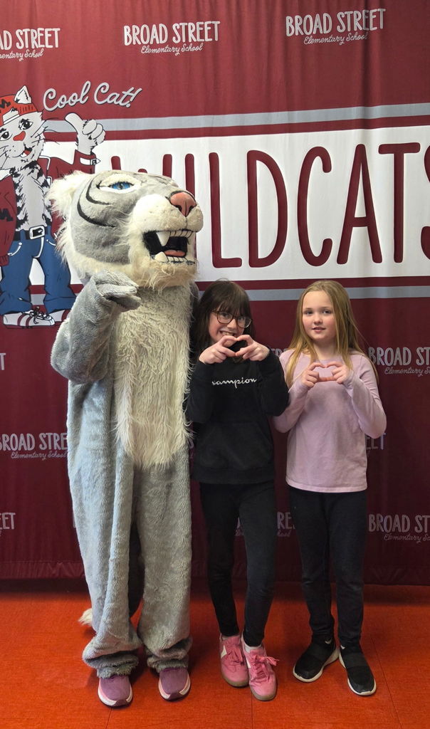 Two young students making hearts with their hands while standing next to a Wildcat mascot.