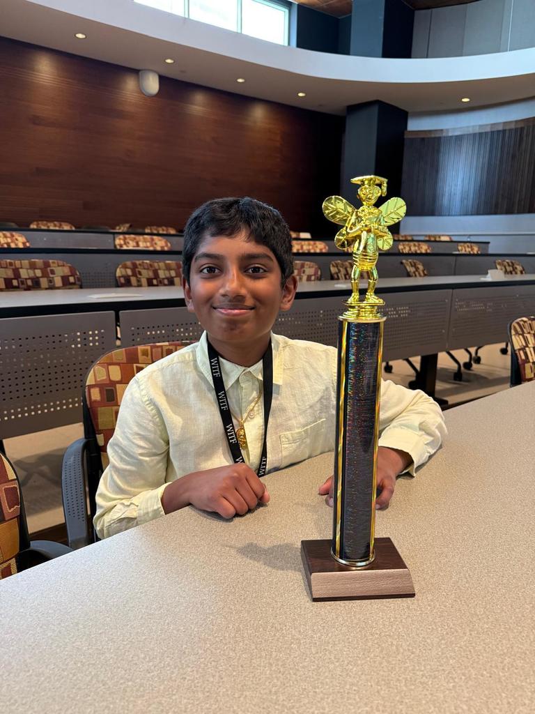 A young student sitting at a table in front of his spelling bee trophy.
