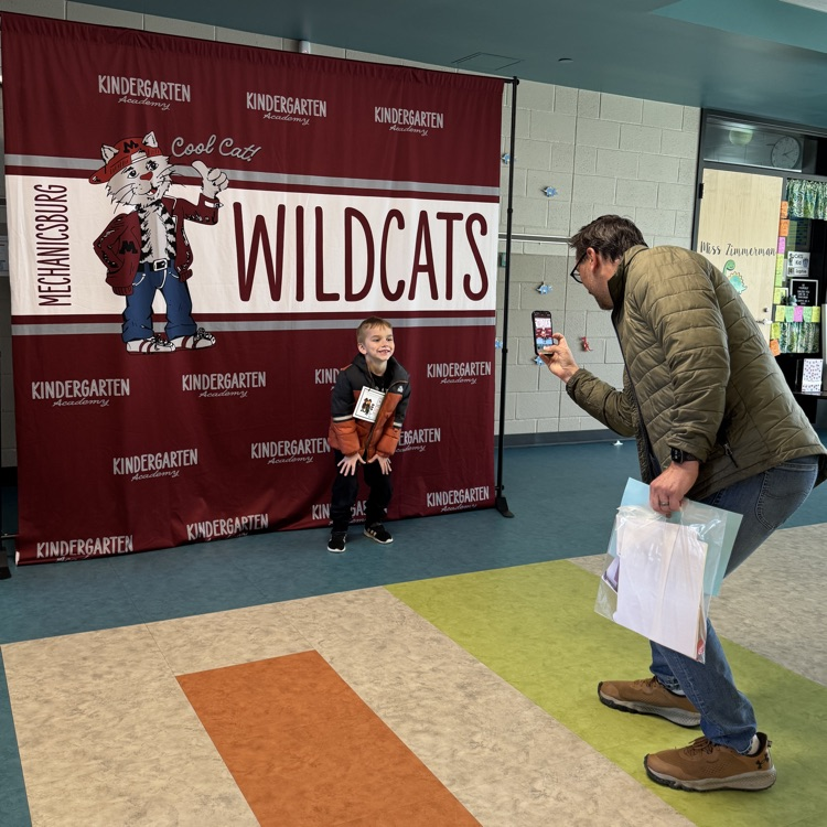 A dad takes a photo of a young boy in front of a wildcat banner.