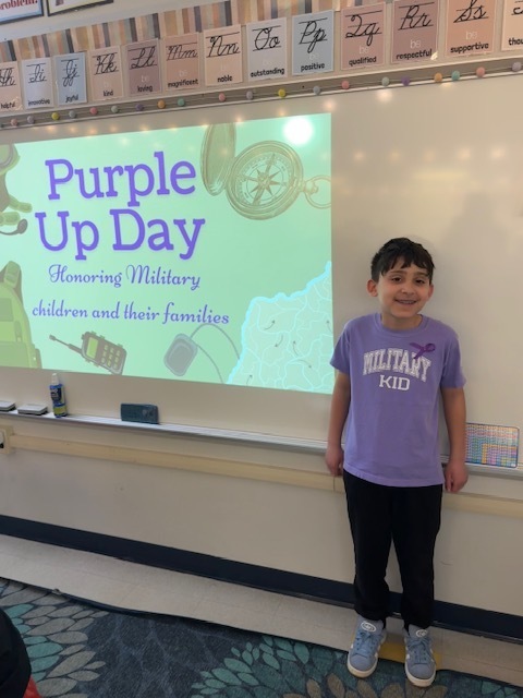 A young student stands next to a display board while wearing a purple "Military Kid" shirt.