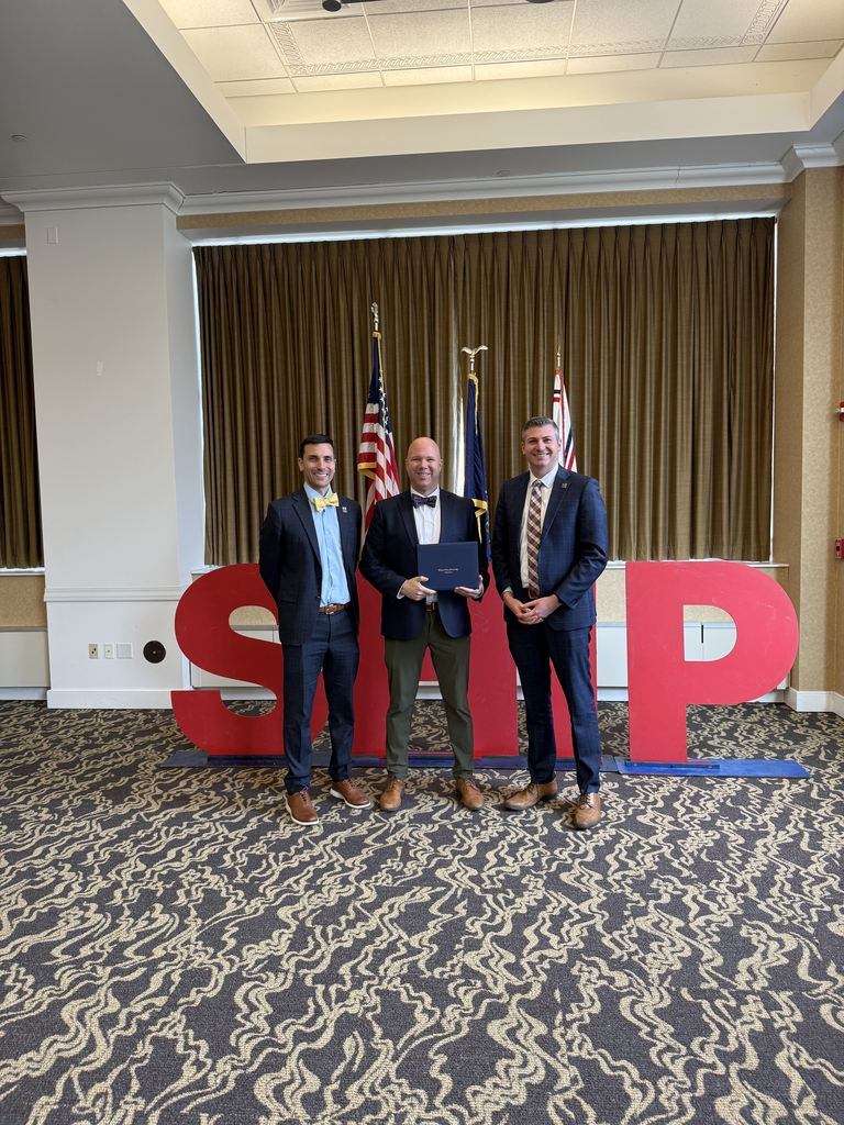 Three men standing in front of big red letters that spell out SHIP as part of an award presentation.