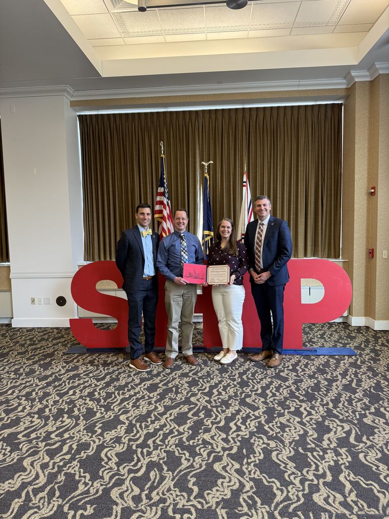 Four adults standing in front of big red letters that spell out SHIP as part of an award presentation.