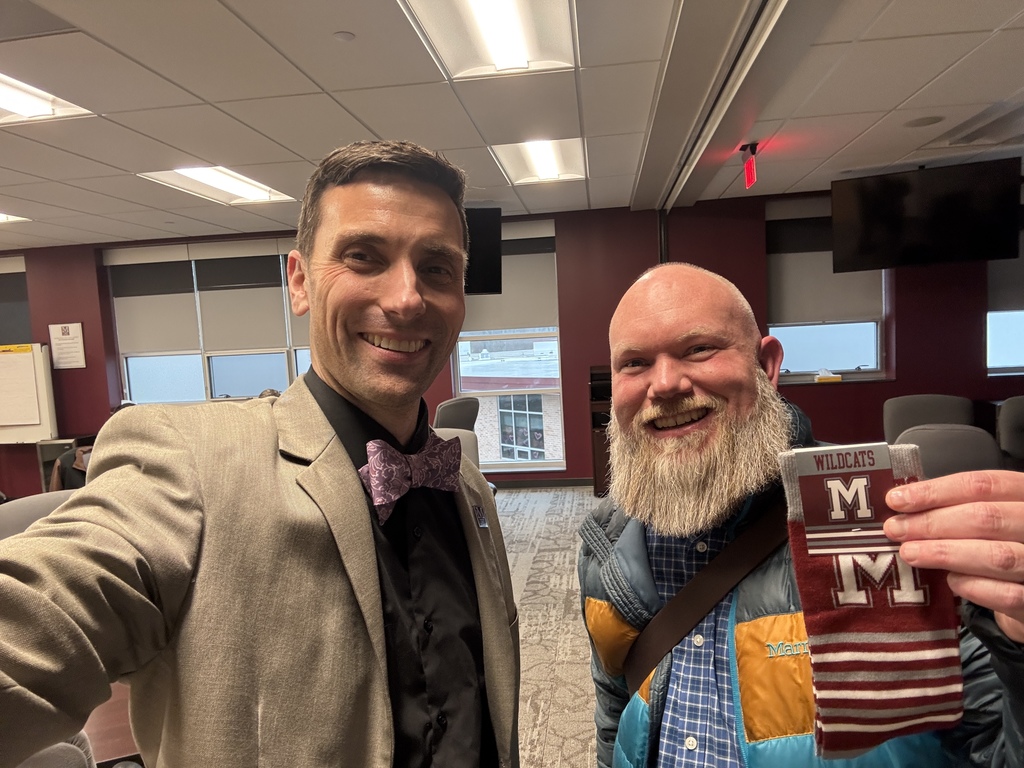 A man in a suit takes a selfie with a man who is holding a pair of branded M socks.