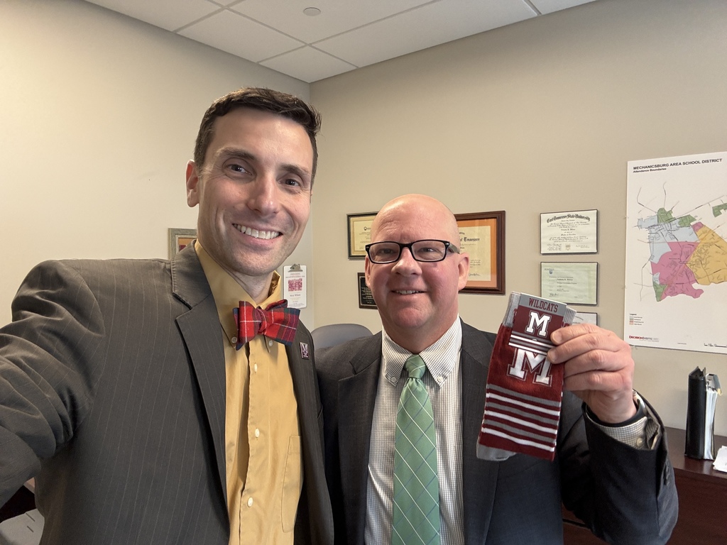 A man in a suit takes a selfie with a man who is holding a pair of branded M socks.