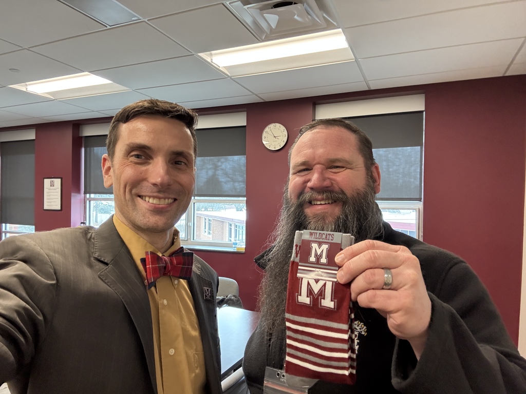 A man in a suit takes a selfie with a man who is holding a pair of branded M socks.
