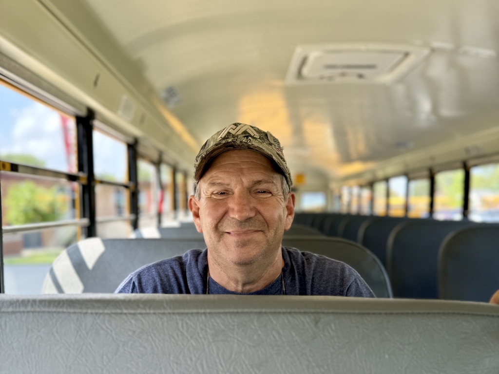 A bus driver sits in a school bus seat and smiles.