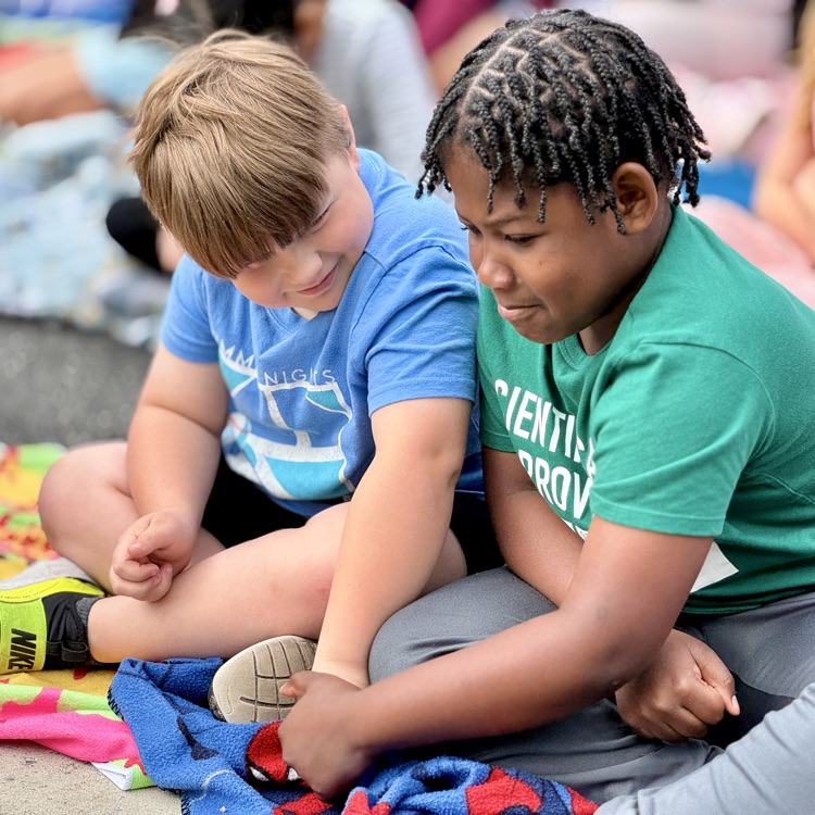 two young students sit on the ground together and smile at each other  