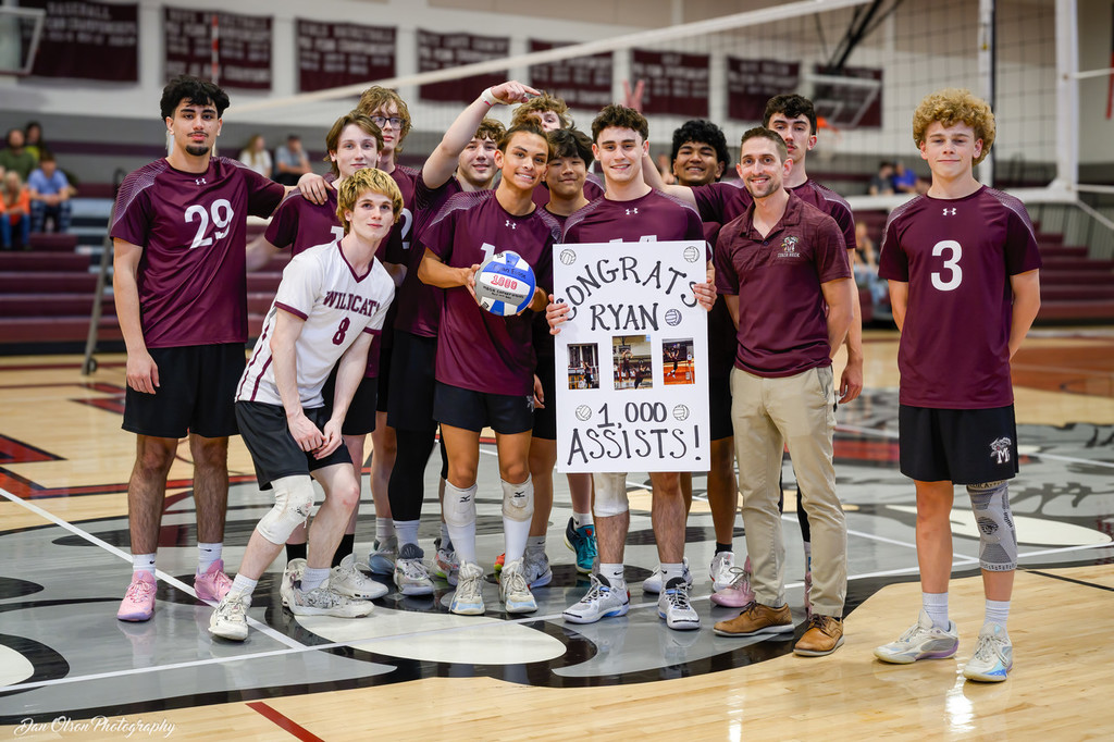 A volleyball team gathers around a teammate who is holding a sign to celebrate is 1000th assist.