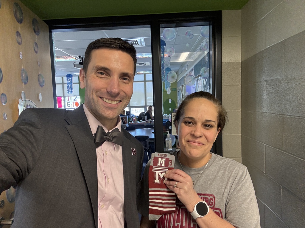 A man a woman standing in front of a school classroom to take a selfie. The woman is holding a pair of branded "M" socks.