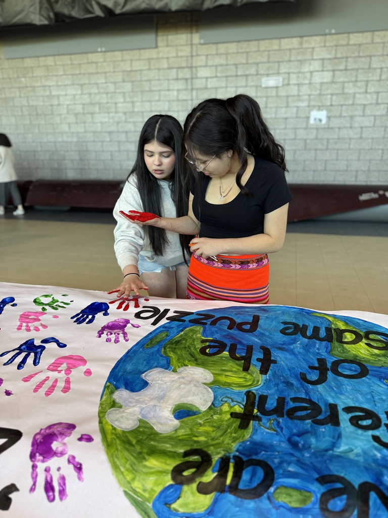 Two students paint their hands to leave a handprint on a colorful canvas.