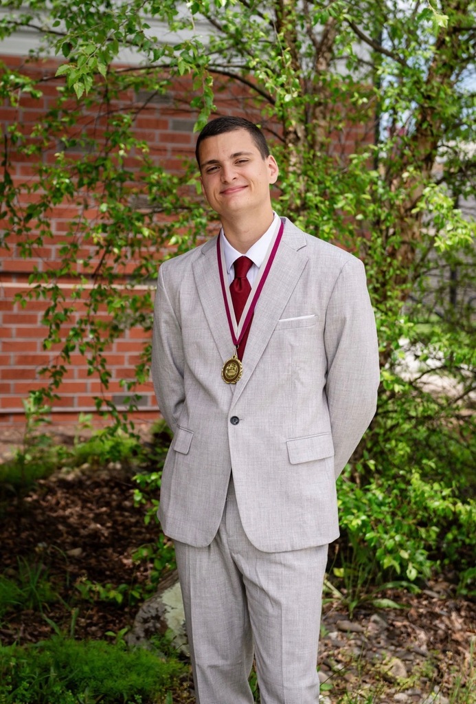 Portrait of a young man in a grey suit standing in front of bushes and wearing a maroon and white medal.
