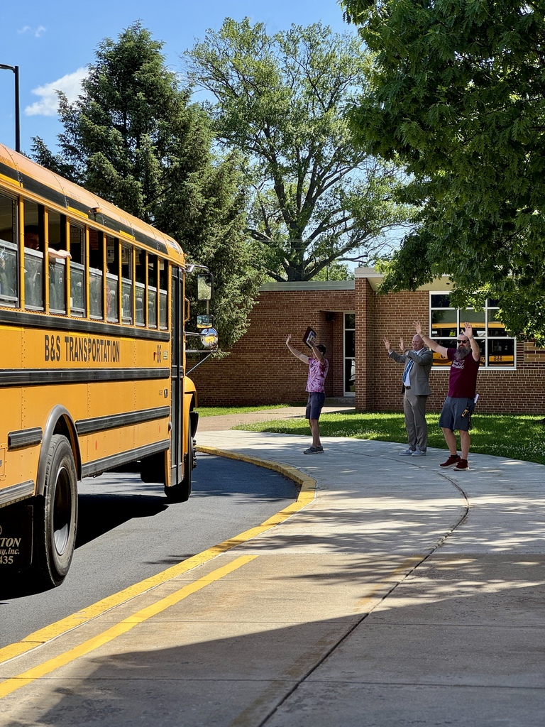 Adults cheer and wave goodbye to a school bus full of students.