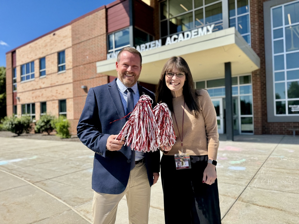 Two adults stand in front of a school with pom poms.