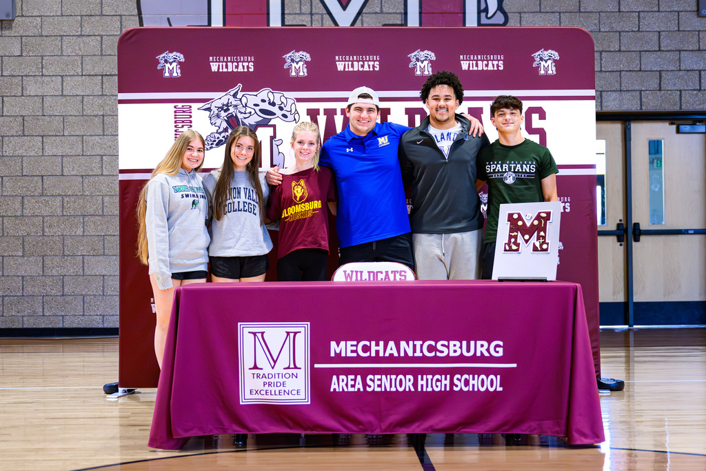 A group of athletes stand behind a table, arm in arm.